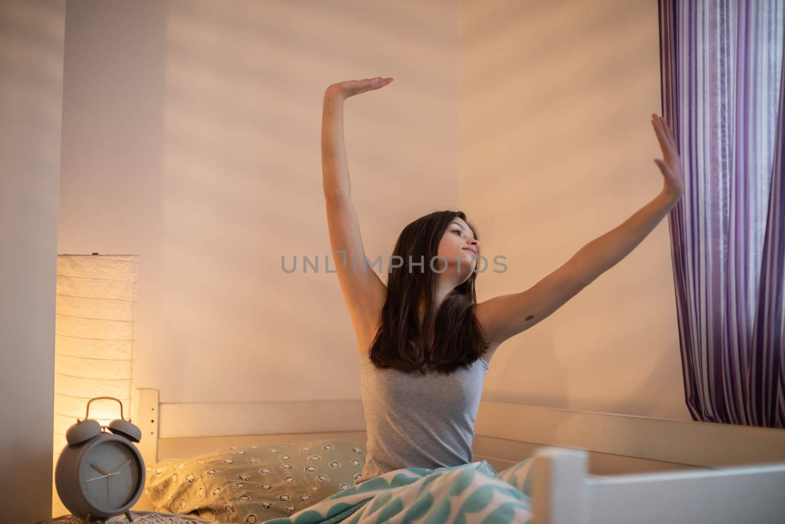 A teenage girl with straight hair stretches in her bedroom on the bed. A large alarm clock stands on the bedside table.