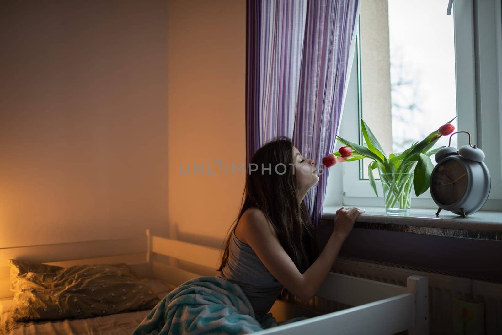 A young woman sniffs the tulips in a vase on the windowsill. Bright bedroom with decorations.