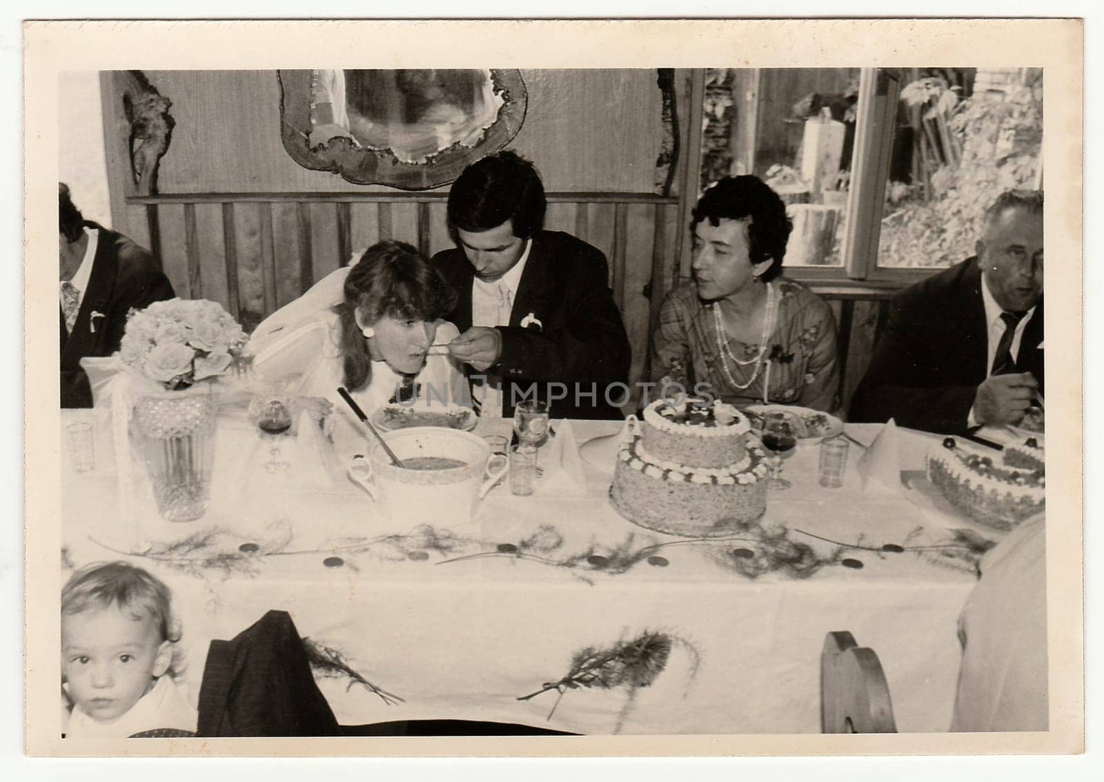 Retro photo shows people during wedding feast. Groom feeds bride. Black and white vintage photography. by roman_nerud