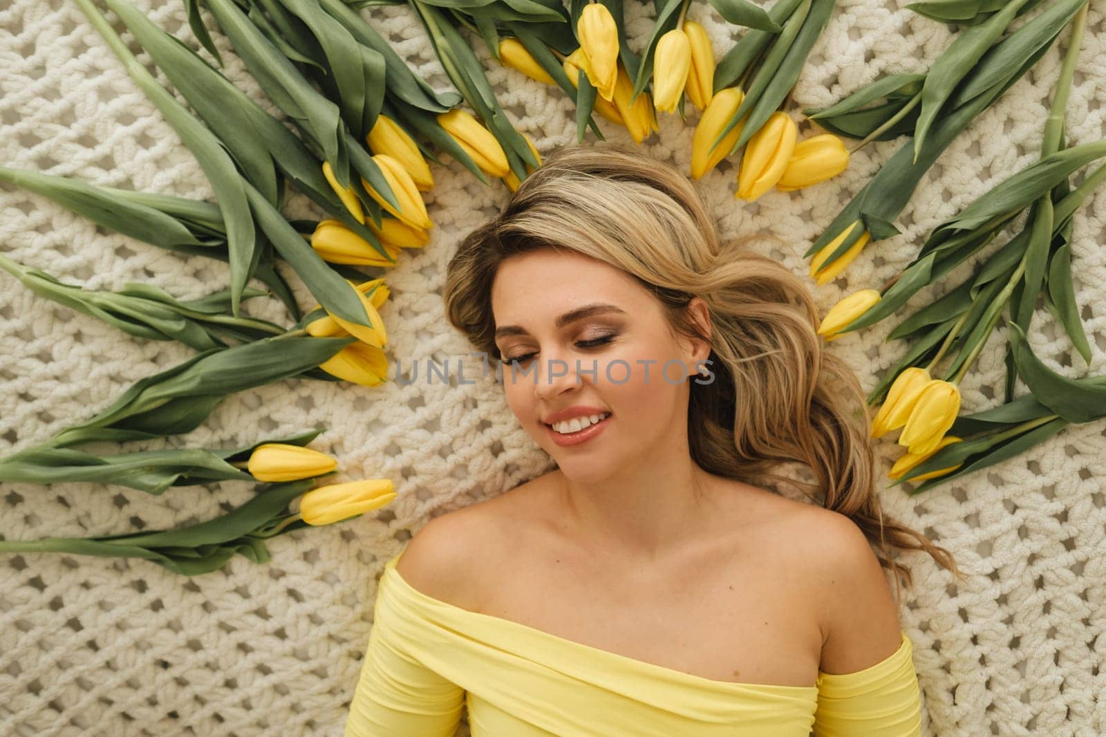 a happy woman in a yellow dress lies in yellow spring tulips on the bed.