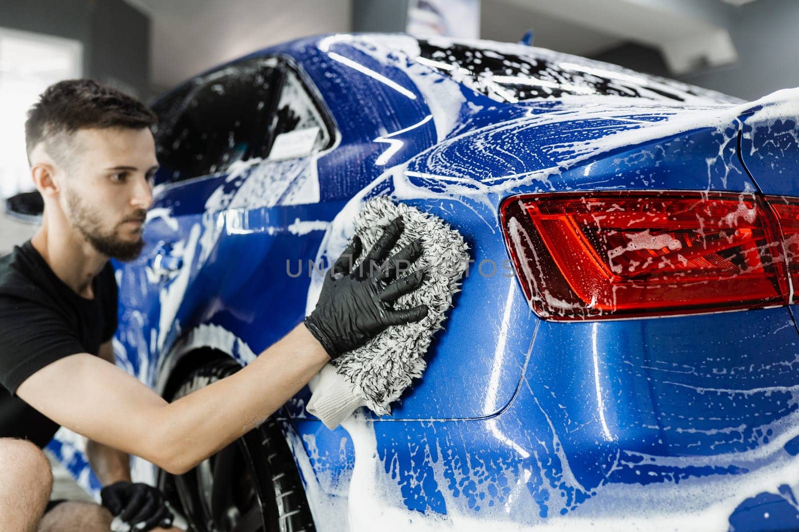 Car washer doing manual foam washing in auto detailing service. Hand washing with microfiber glove with foam car body in garage. by Rabizo