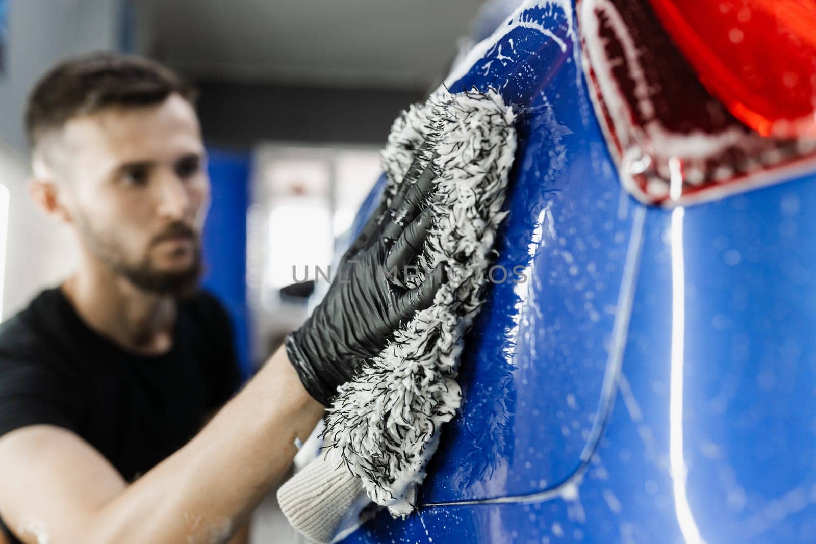 Car washer doing manual foam washing in auto detailing service. Hand washing with microfiber glove with foam car body in garage. by Rabizo