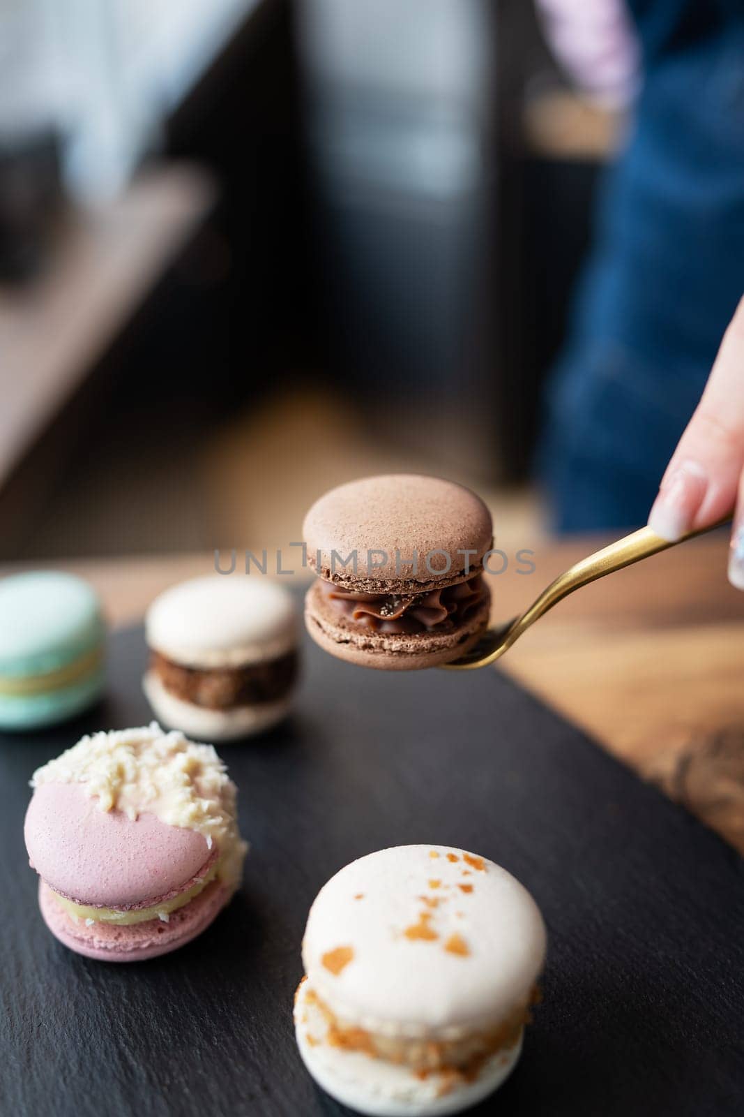 A delicious macaron dessert - a French sweet delicacy lies on a concrete black stand, and a girl holds a candy with a fork. Vertical photo. by sfinks