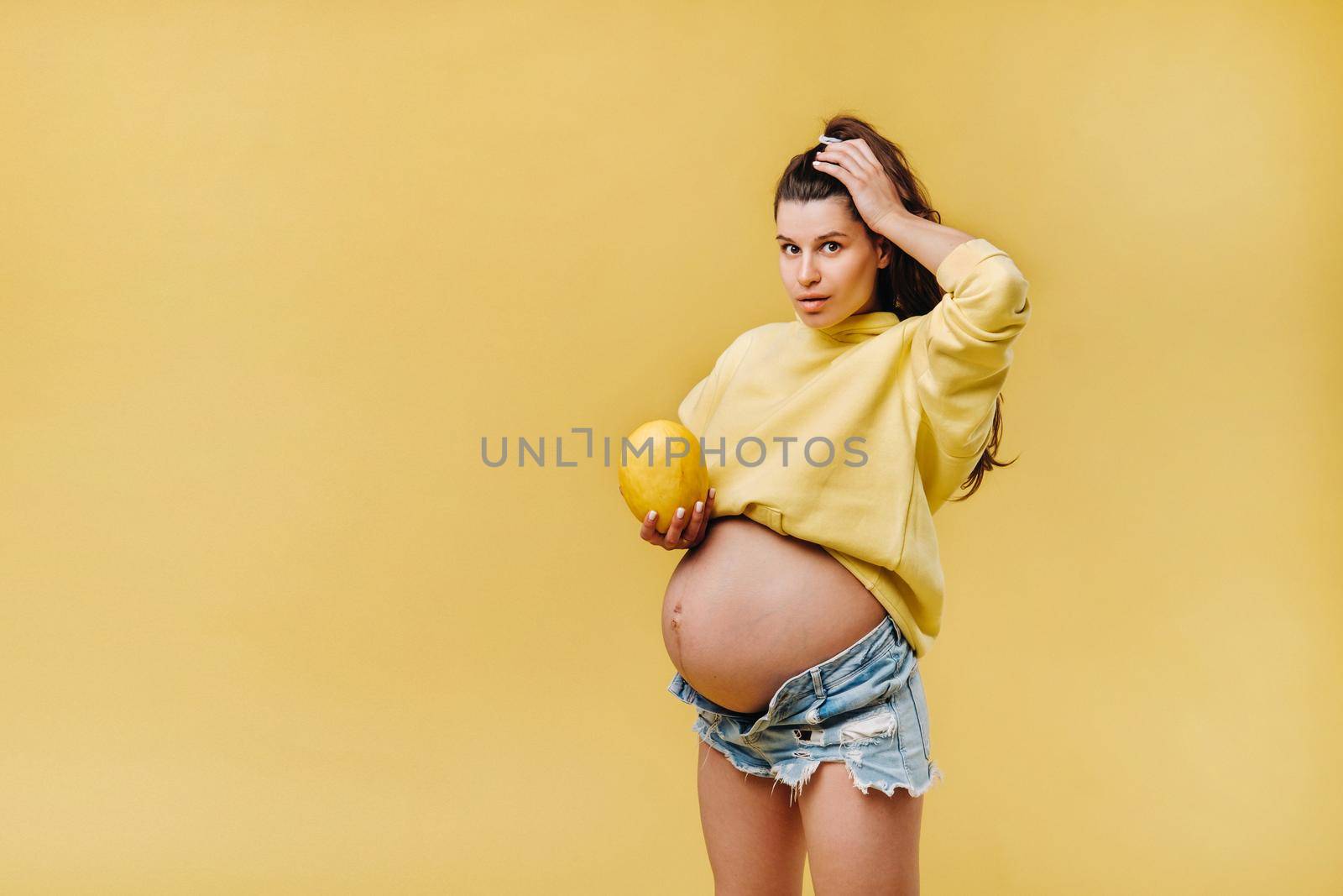 a pregnant girl with a yellow jacket stands with a melon in her hands on a yellow isolated background.