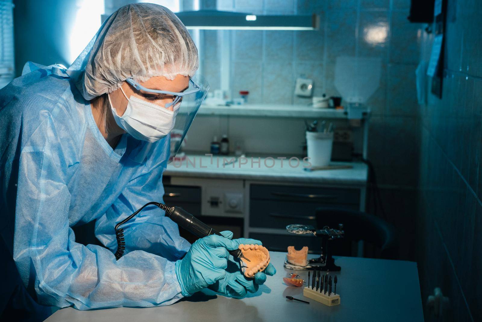 A masked and gloved dental technician works on a prosthetic tooth in his lab by Lobachad