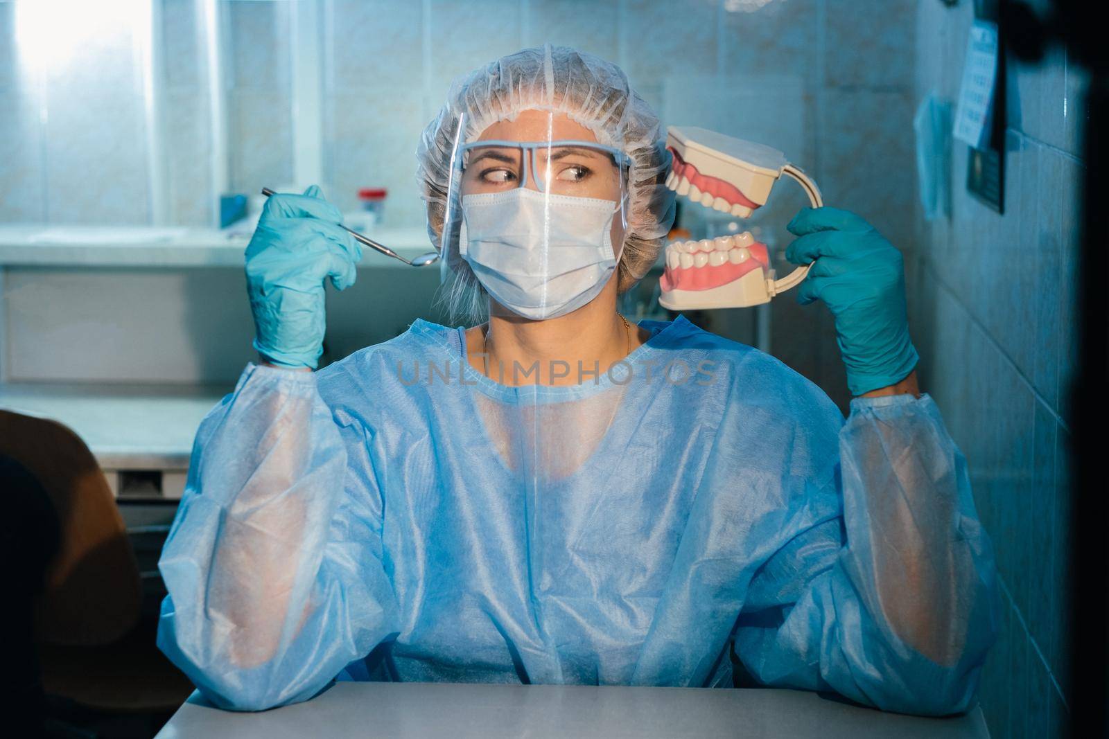 A dental doctor wearing blue gloves and a mask holds a dental model of the upper and lower jaws and a dental mirror.