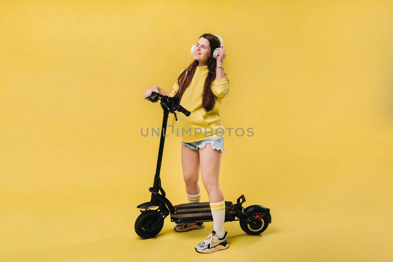 Pregnant girl in yellow clothes and headphones on an electric scooter on an isolated yellow background by Lobachad