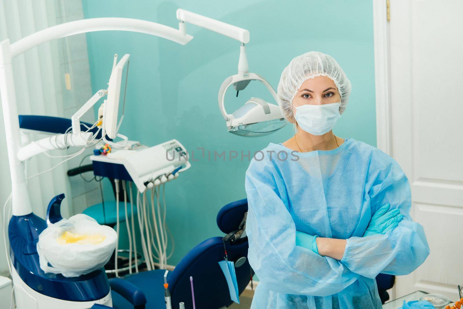 a female dentist wearing a medical mask and rubber gloves poses for the camera and folds her arms in her office by Lobachad