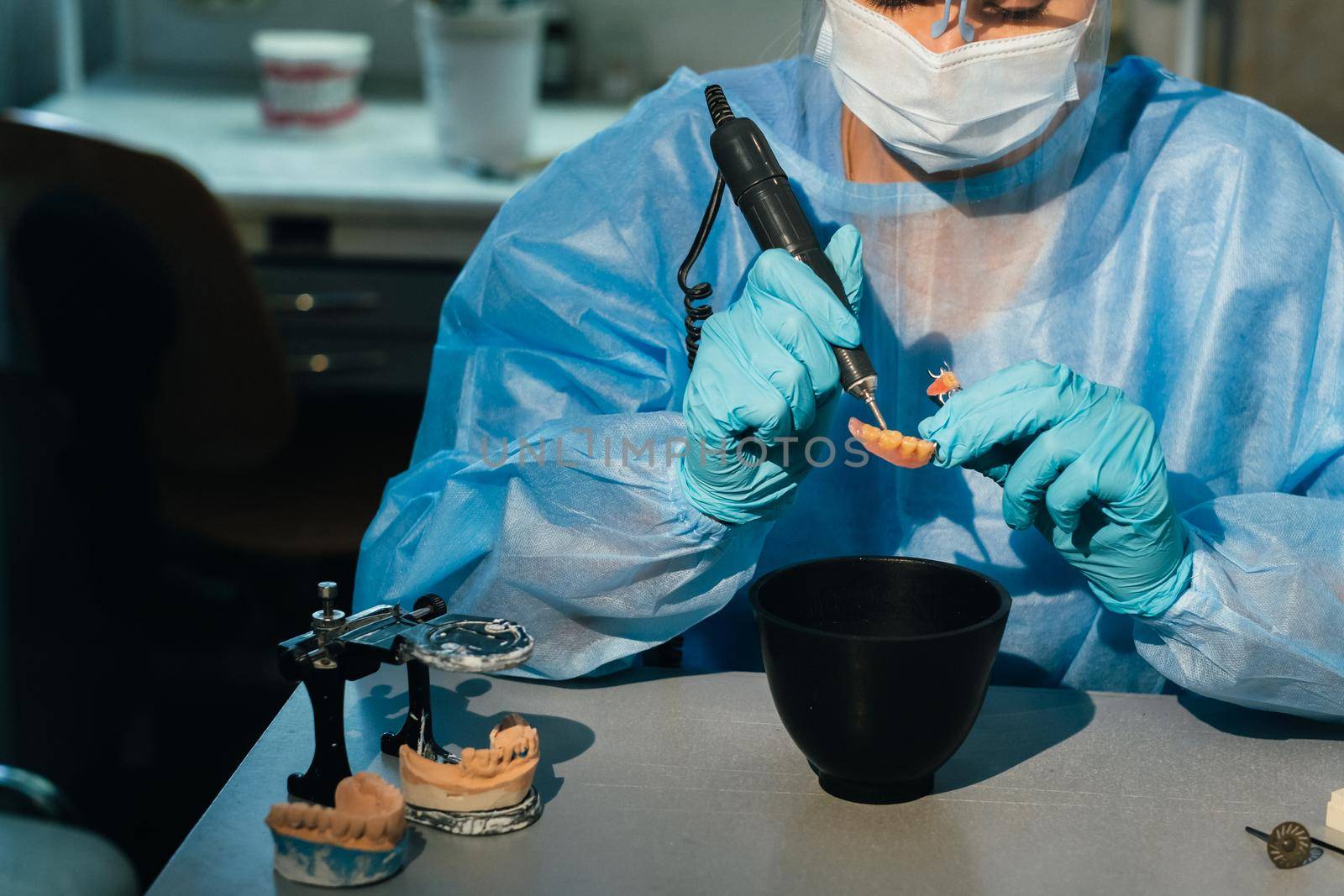 A masked and gloved dental technician works on a prosthetic tooth in his lab.