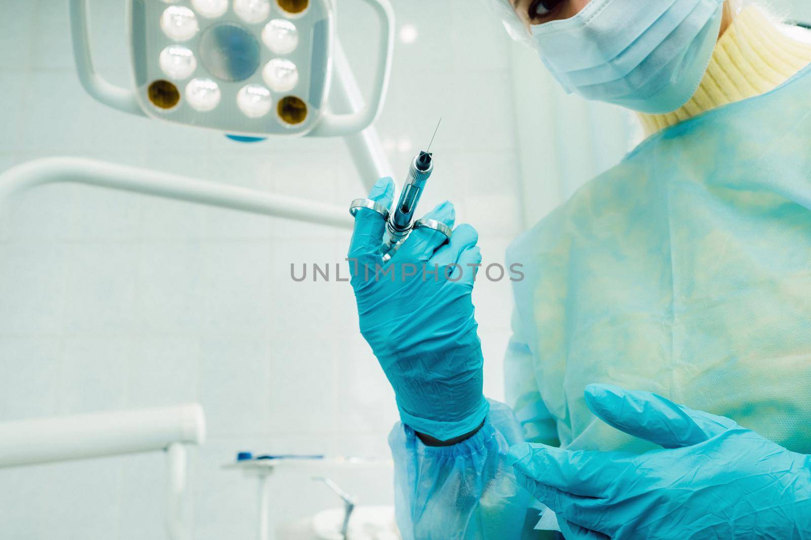 A masked dentist holds an injection syringe for a patient in the office by Lobachad