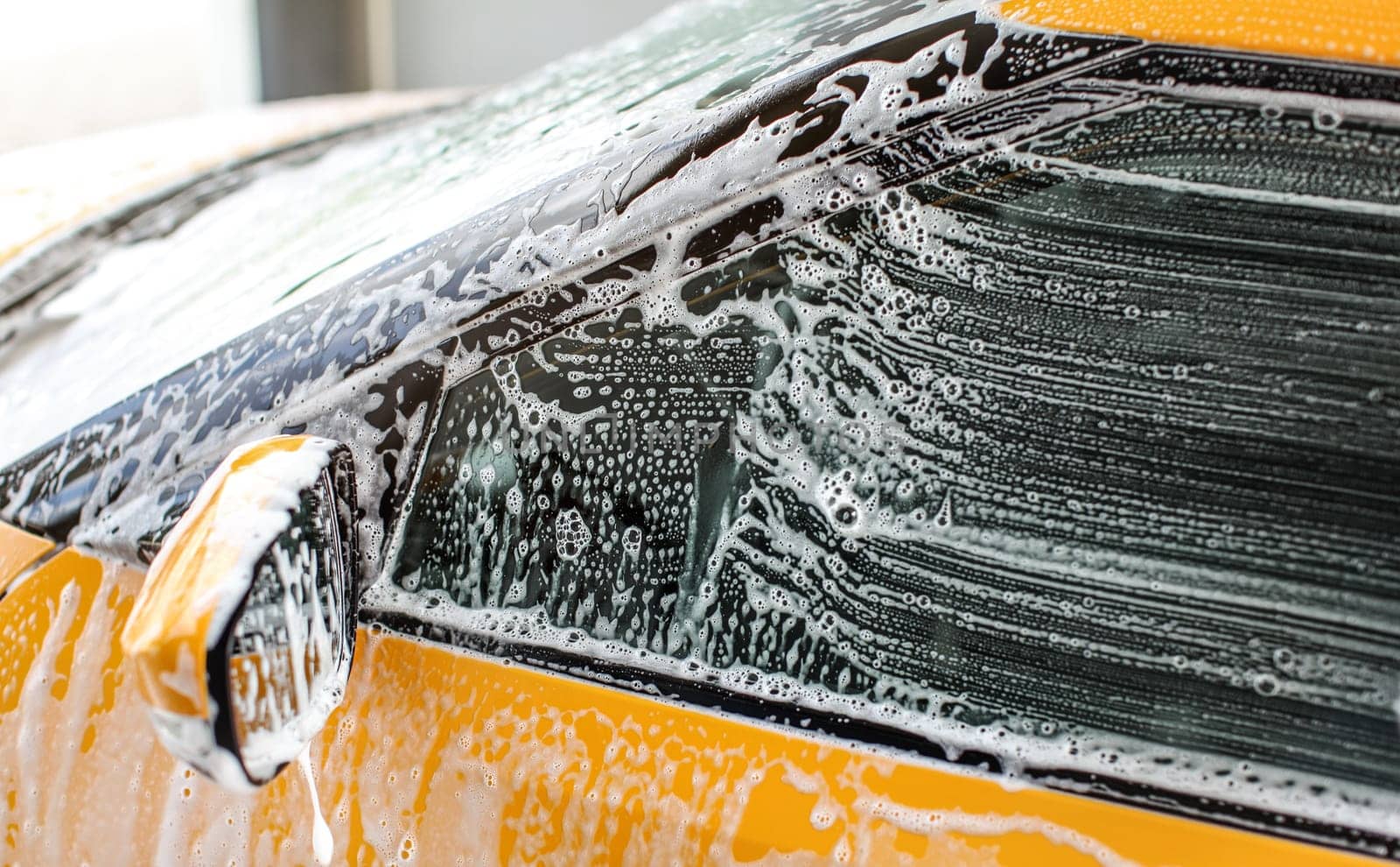 Side window and mirror of yellow car washed in carwash. Strokes from brush visible on glass in white shampoo and foam.
