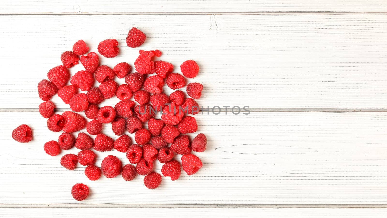 Tabletop view, pile of freshly picked raspberries spilled on white boards desk. Space for text on the right.