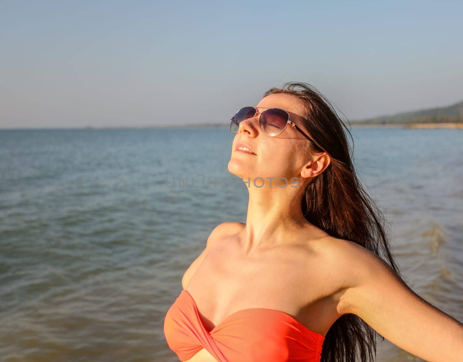 Young woman in red bikini and sunglasses, arms spread, looking into and enjoying afternoon sun, sea behind. Detail on head and top of her body.