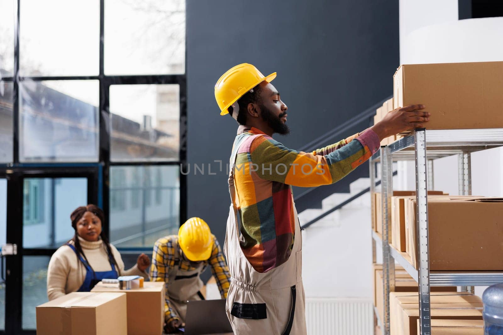Warehouse worker in hard hat taking carton from metal shelf while standing on ladder in storage room. African american package handler reaching for cardboard box to prepare customer order