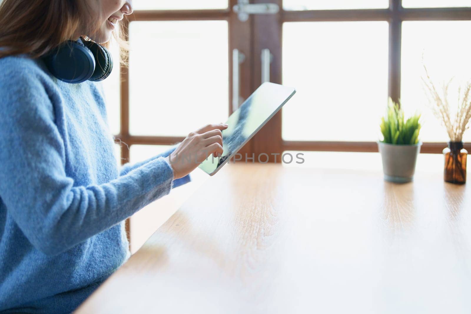 Portrait of a teenage Asian woman using a tablet, wearing headphones to study online via video conferencing on a wooden desk in library.