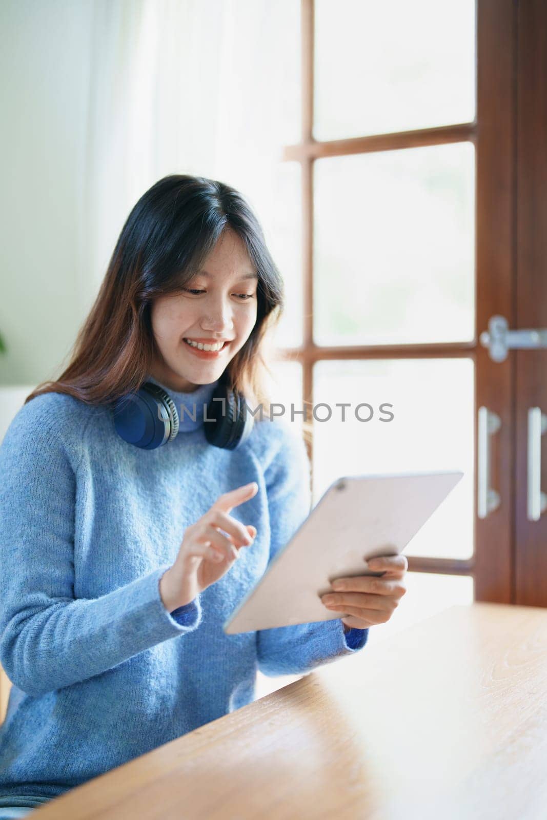 Portrait of a teenage Asian woman using a tablet, wearing headphones to study online via video conferencing on a wooden desk in library by Manastrong