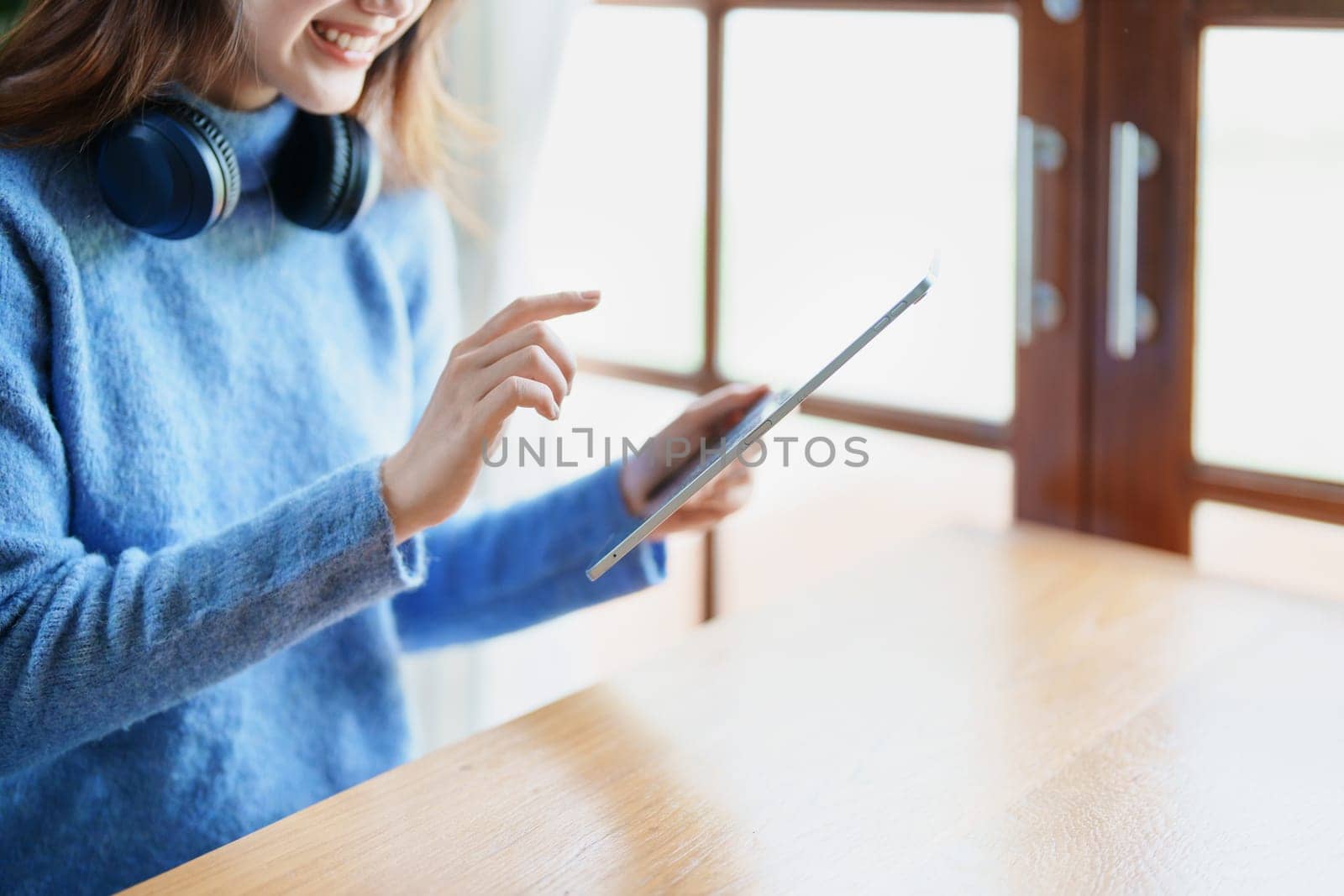 Portrait of a teenage Asian woman using a tablet, wearing headphones to study online via video conferencing on a wooden desk in library by Manastrong