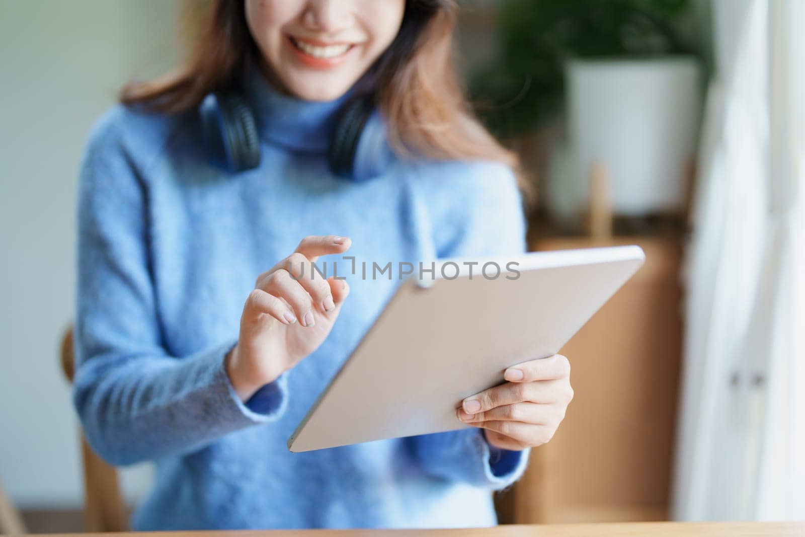 Portrait of a teenage Asian woman using a tablet, wearing headphones to study online via video conferencing on a wooden desk in library by Manastrong