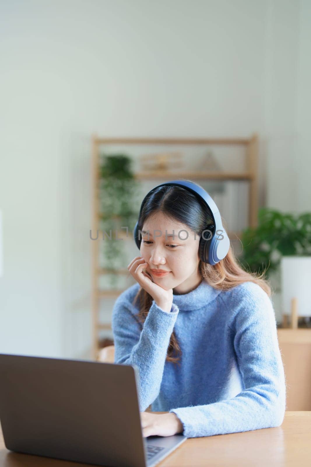Portrait of a teenage Asian woman using a computer, wearing headphones to study online via video conferencing on a wooden desk at home by Manastrong