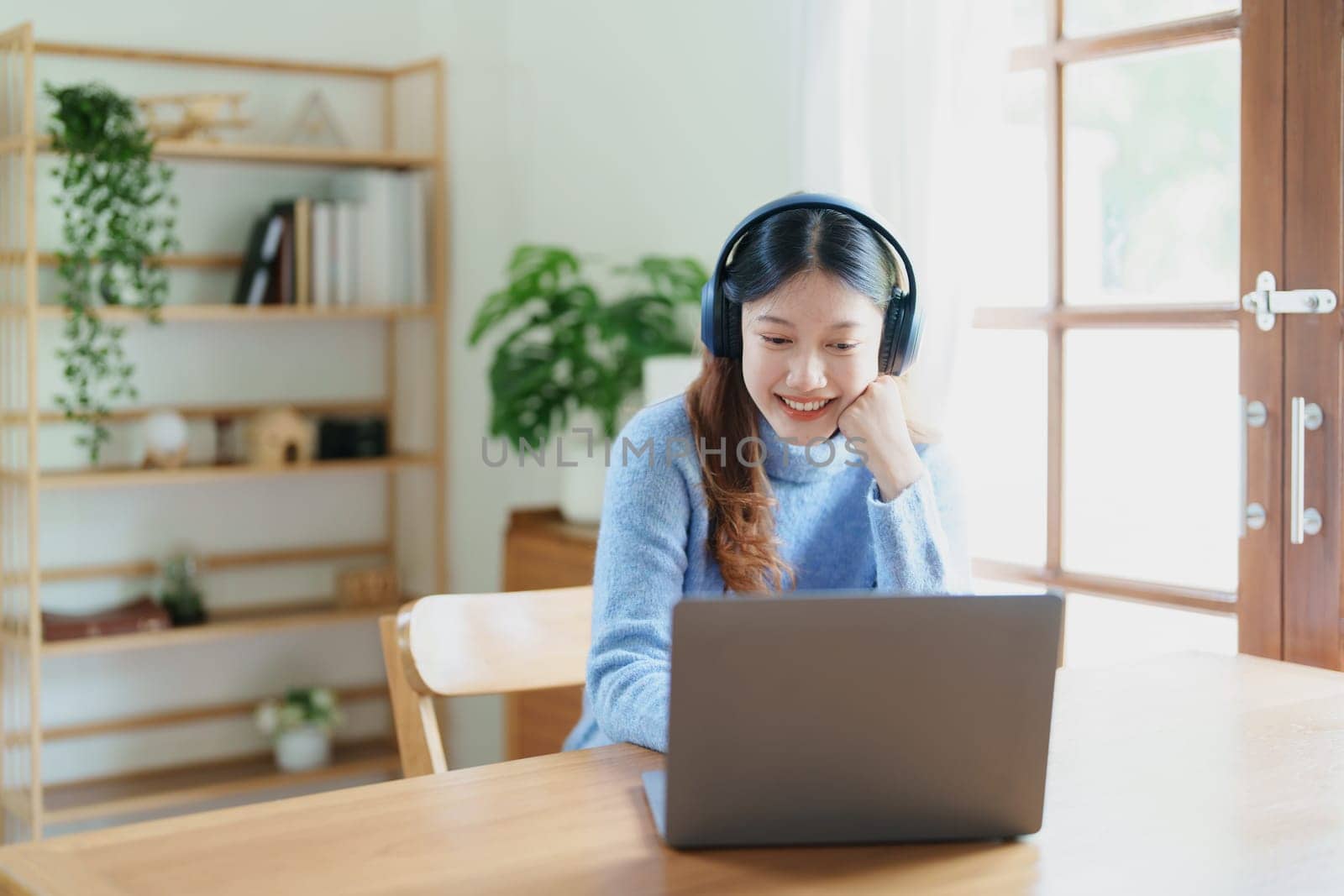Portrait of a teenage Asian woman using a computer, wearing headphones to study online via video conferencing on a wooden desk at home.