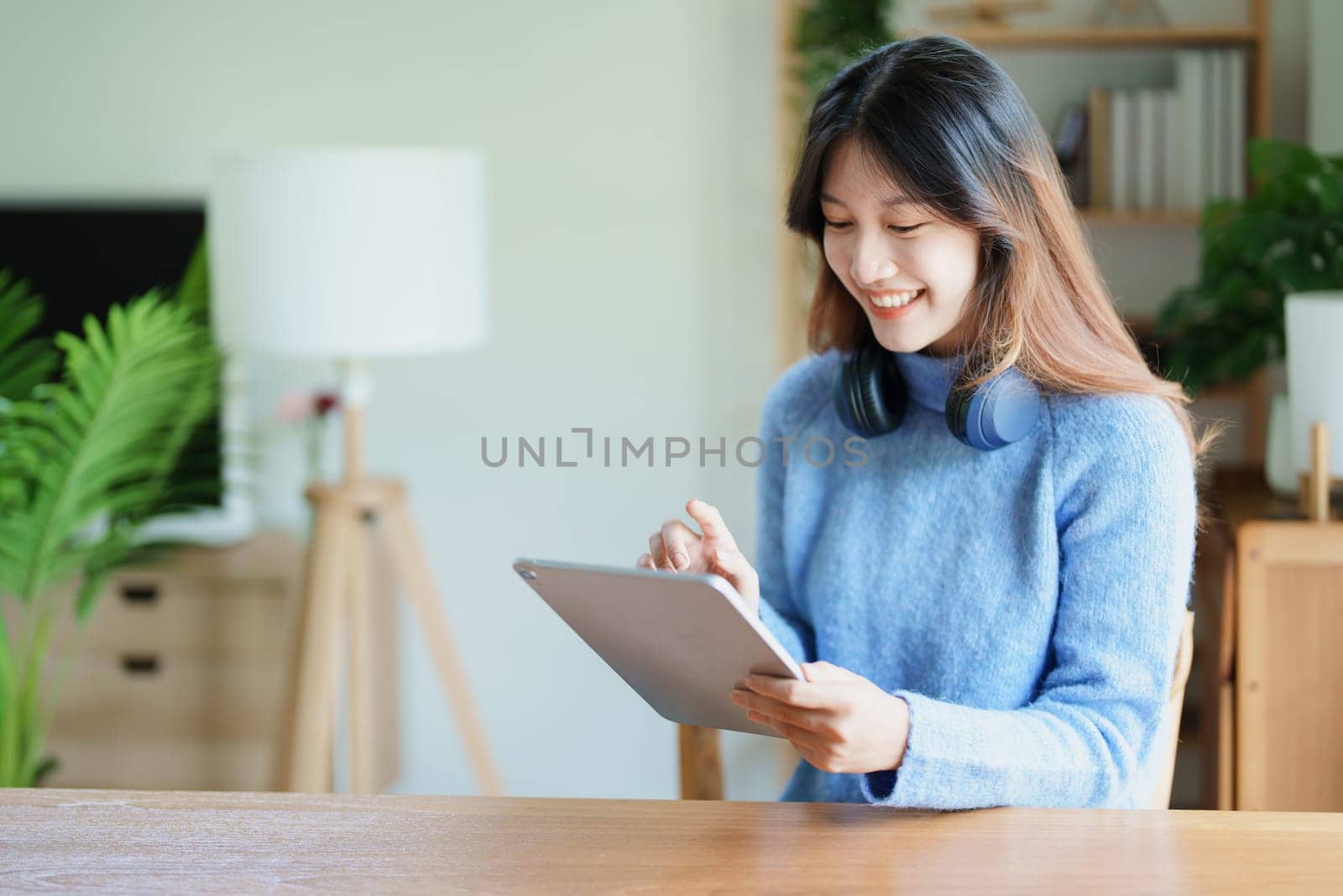 Portrait of a teenage Asian woman using a tablet, wearing headphones to study online via video conferencing on a wooden desk in library.