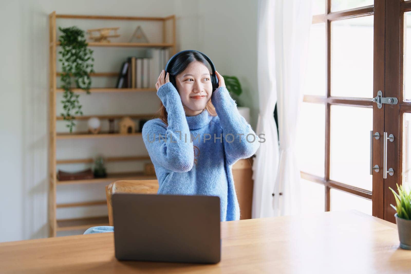 Portrait of a teenage Asian woman using a computer, wearing headphones to study online via video conferencing on a wooden desk at home.