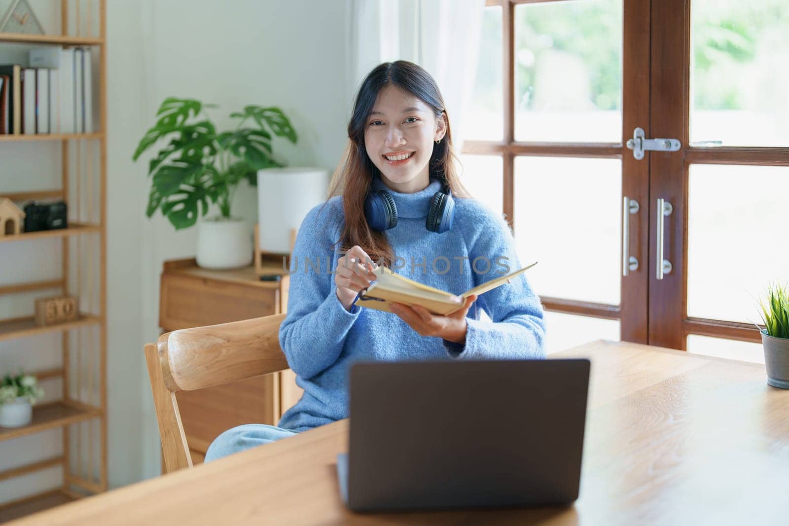 Portrait of a teenage Asian woman using a computer, wearing headphones and using a notebook to study online via video conferencing on a wooden desk in library by Manastrong