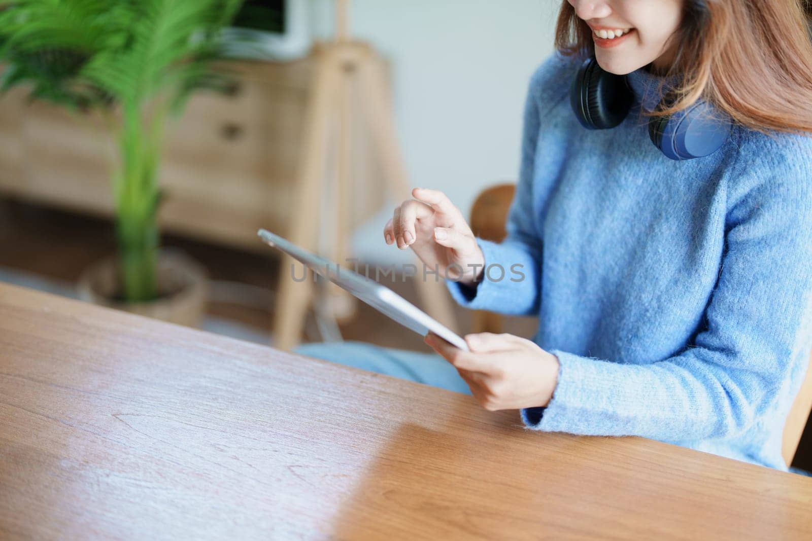 Portrait of a teenage Asian woman using a tablet, wearing headphones to study online via video conferencing on a wooden desk in library.