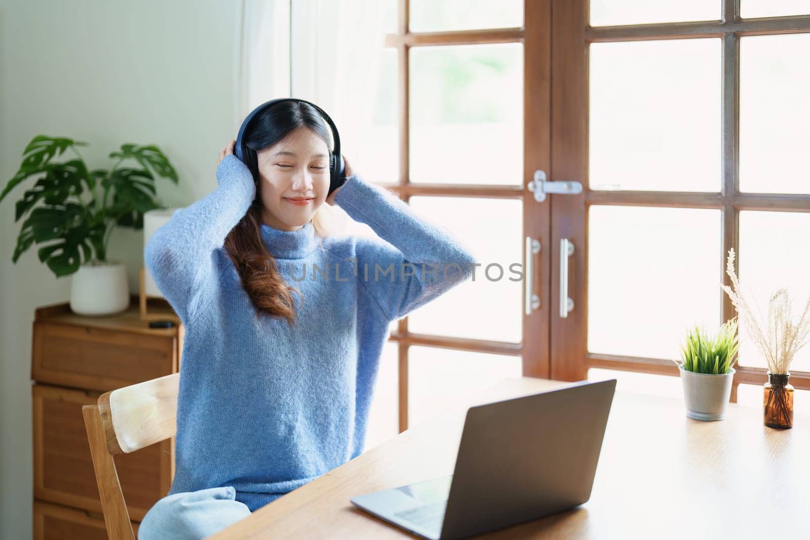 Portrait of a teenage Asian woman using a computer, wearing headphones to study online via video conferencing on a wooden desk at home by Manastrong