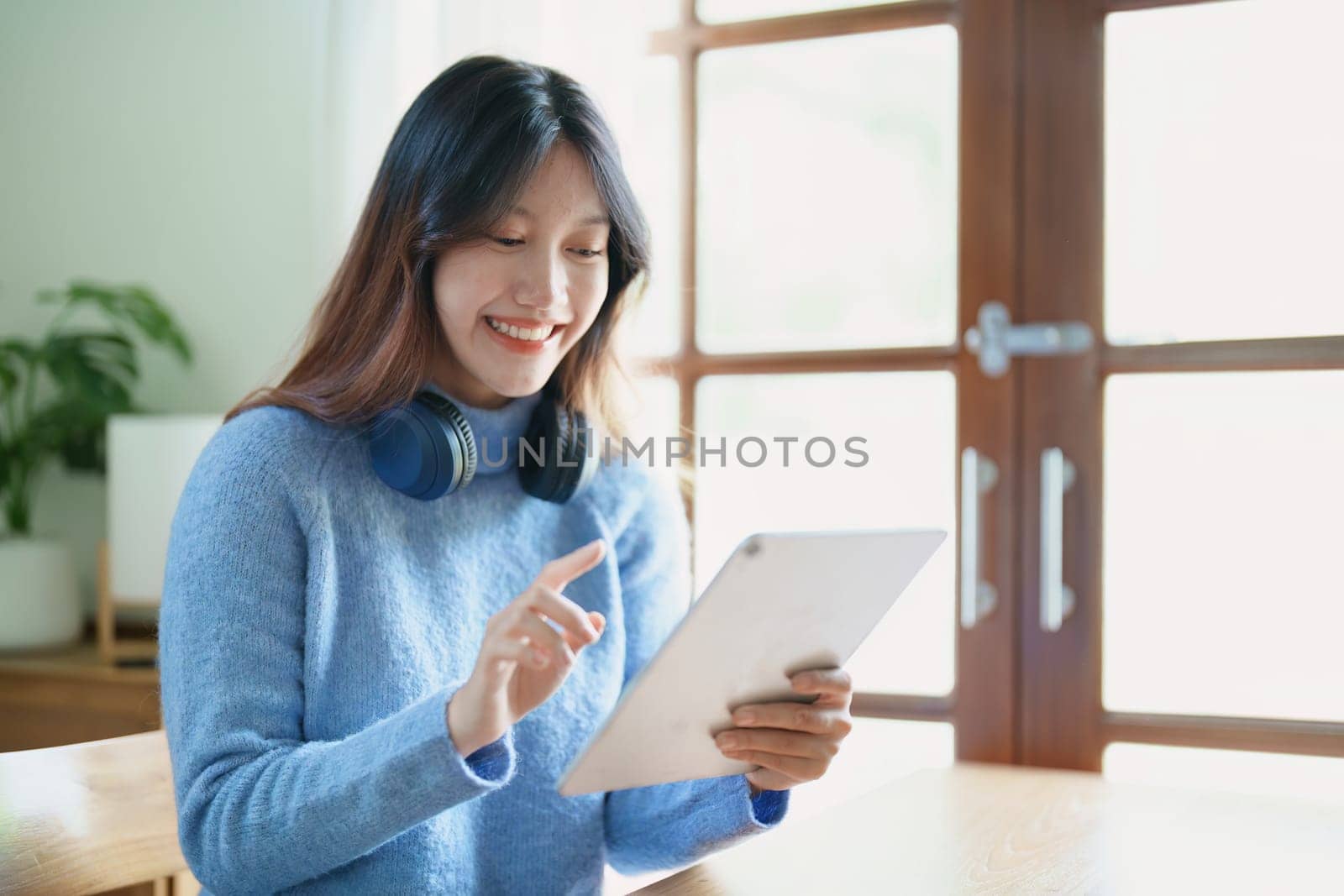 Portrait of a teenage Asian woman using a tablet, wearing headphones to study online via video conferencing on a wooden desk in library by Manastrong