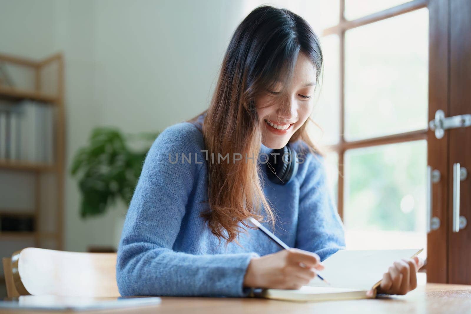 Portrait of a teenage Asian woman using a notebook to study online via video conferencing on a wooden desk at home by Manastrong