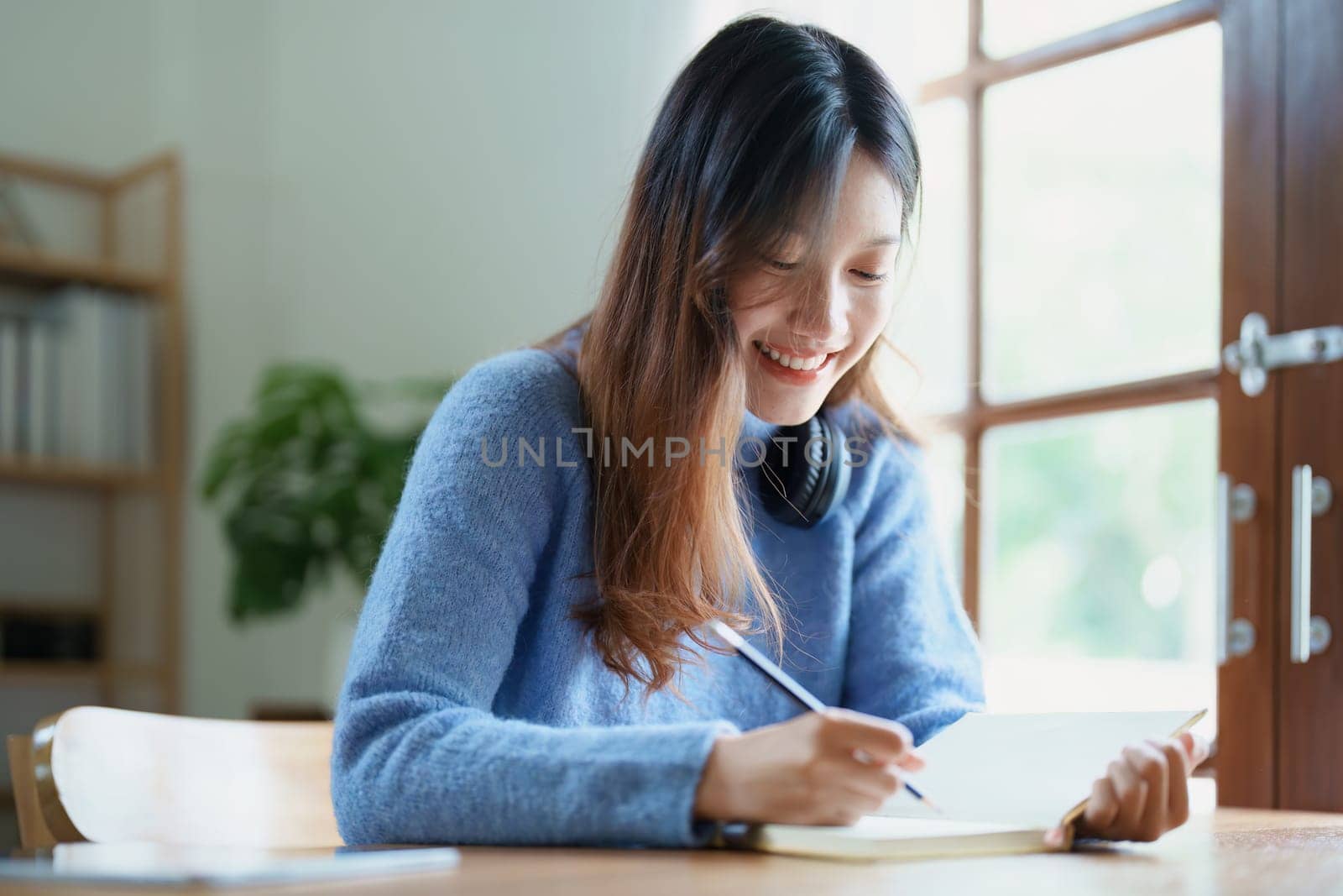 Portrait of a teenage Asian woman using a notebook to study online via video conferencing on a wooden desk at home.