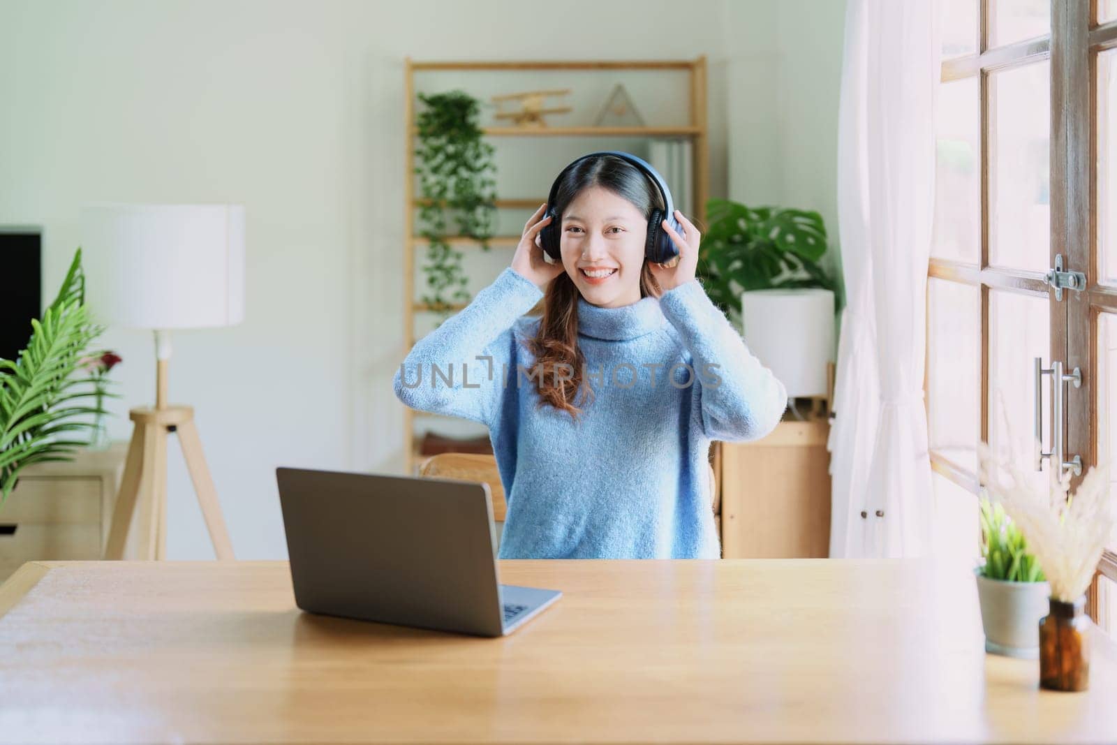 Portrait of a teenage Asian woman using a computer, wearing headphones to study online via video conferencing on a wooden desk at home by Manastrong