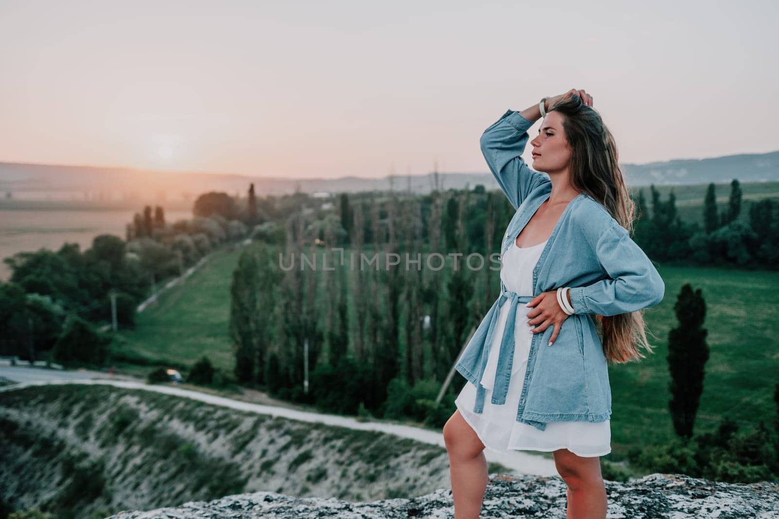 Happy woman standing with her back on the sunset in nature in summer with open hands. Romantic beautiful bride in white boho dress posing with mountains on sunset by panophotograph