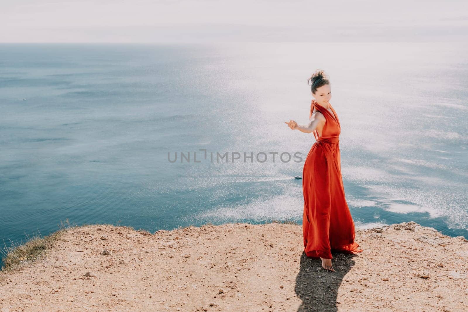 Side view a Young beautiful sensual woman in a red long dress posing on a rock high above the sea during sunrise. Girl on the nature on blue sky background. Fashion photo.