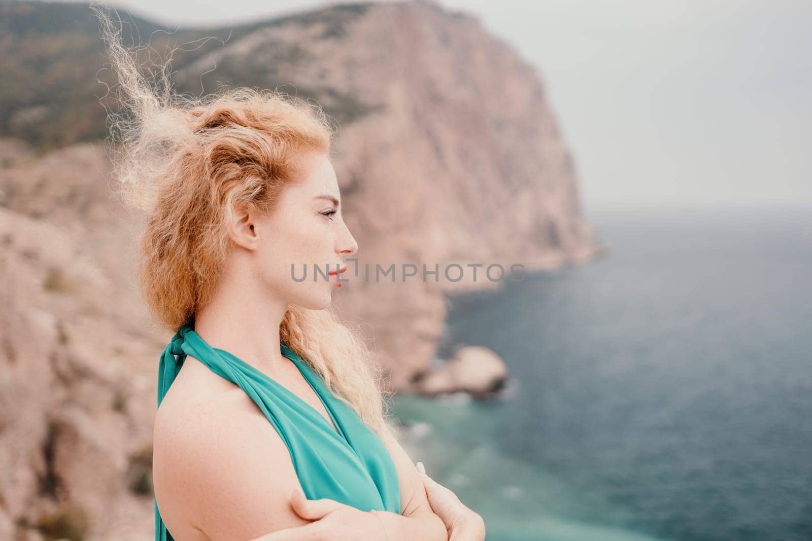 Redhead woman portrait. Curly redhead young caucasian woman with freckles looking at camera and smiling. Close up portrait cute woman in a mint long dress posing on a volcanic rock high above the sea by panophotograph