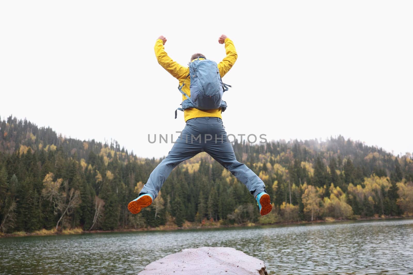 Back view of happy male traveler with backpack jumping near forest lake. Achieving goals while traveling concept