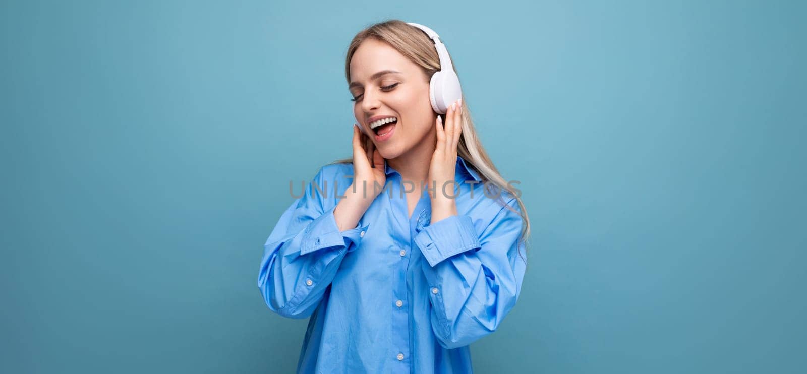 shy blonde girl in a casual shirt listens to songs in big headphones and sings along on a blue background with empty space.