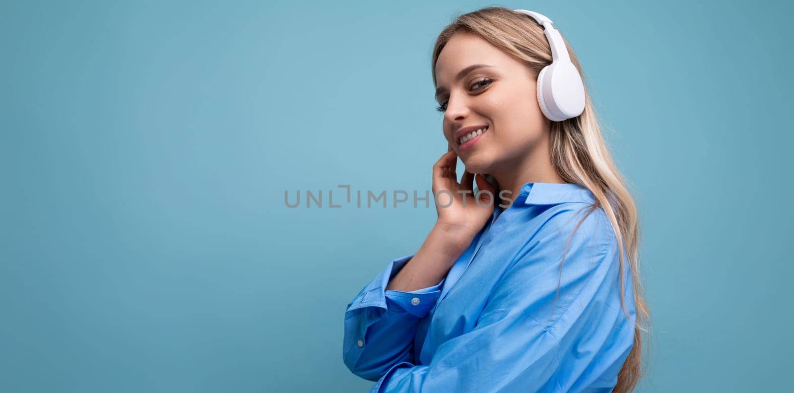 horizontal photo of an attractive girl in wireless large headphones listening with pleasure to music from a playlist on a blue background.