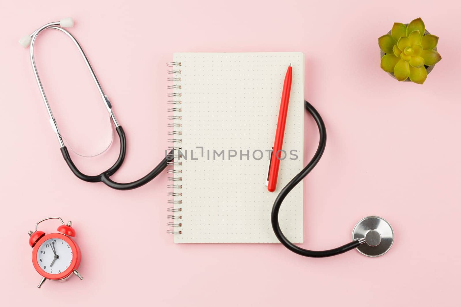 Spiral notepad with a pen on a pink background. Top view. Stethoscope with alarm clock on the doctor table. Flat lay.