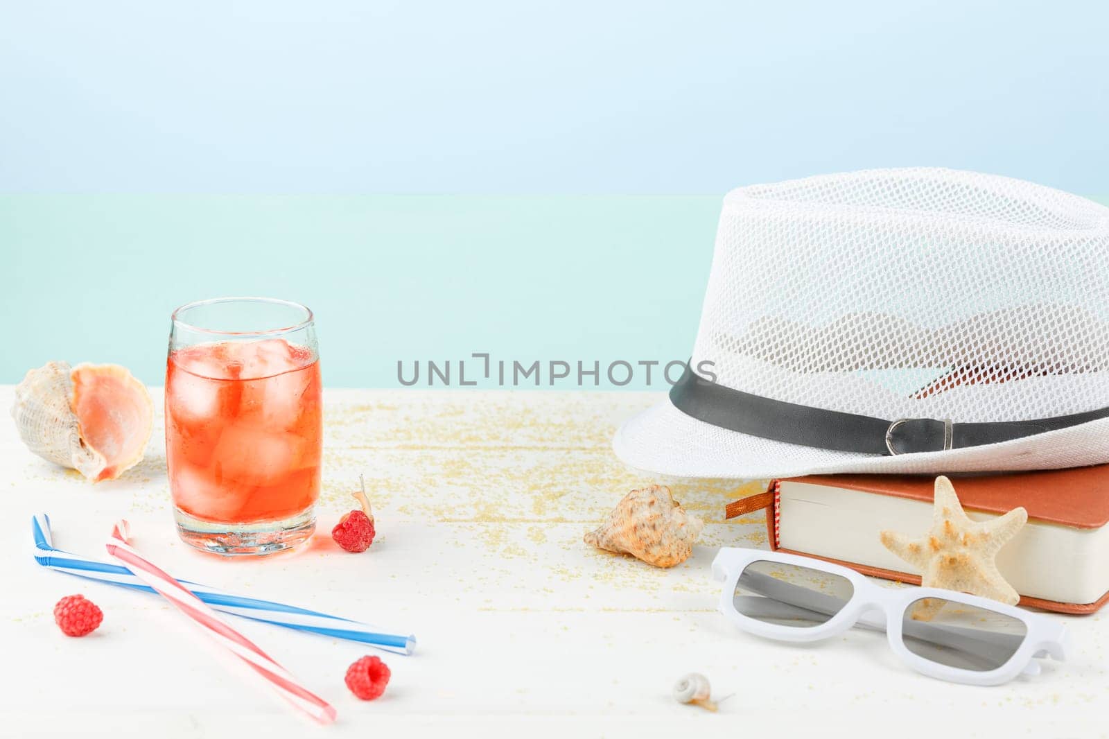Summertime relax. Summer accessories on a white wooden table. Sea vacation concept. Cocktail with ice and red raspberries. Seashells with starfish and snails.