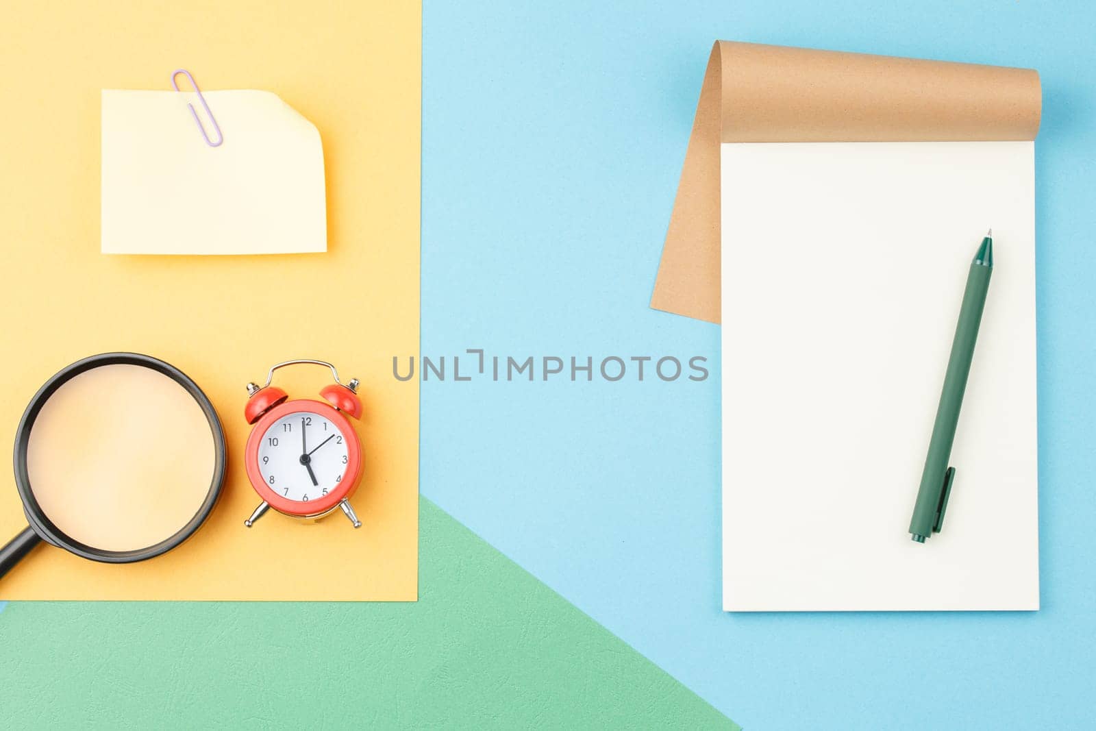 Open notebook with a pen, alarm clock, magnifying glass and paper for notes with a paper clip on a colored paper background. Top view. Office concept.