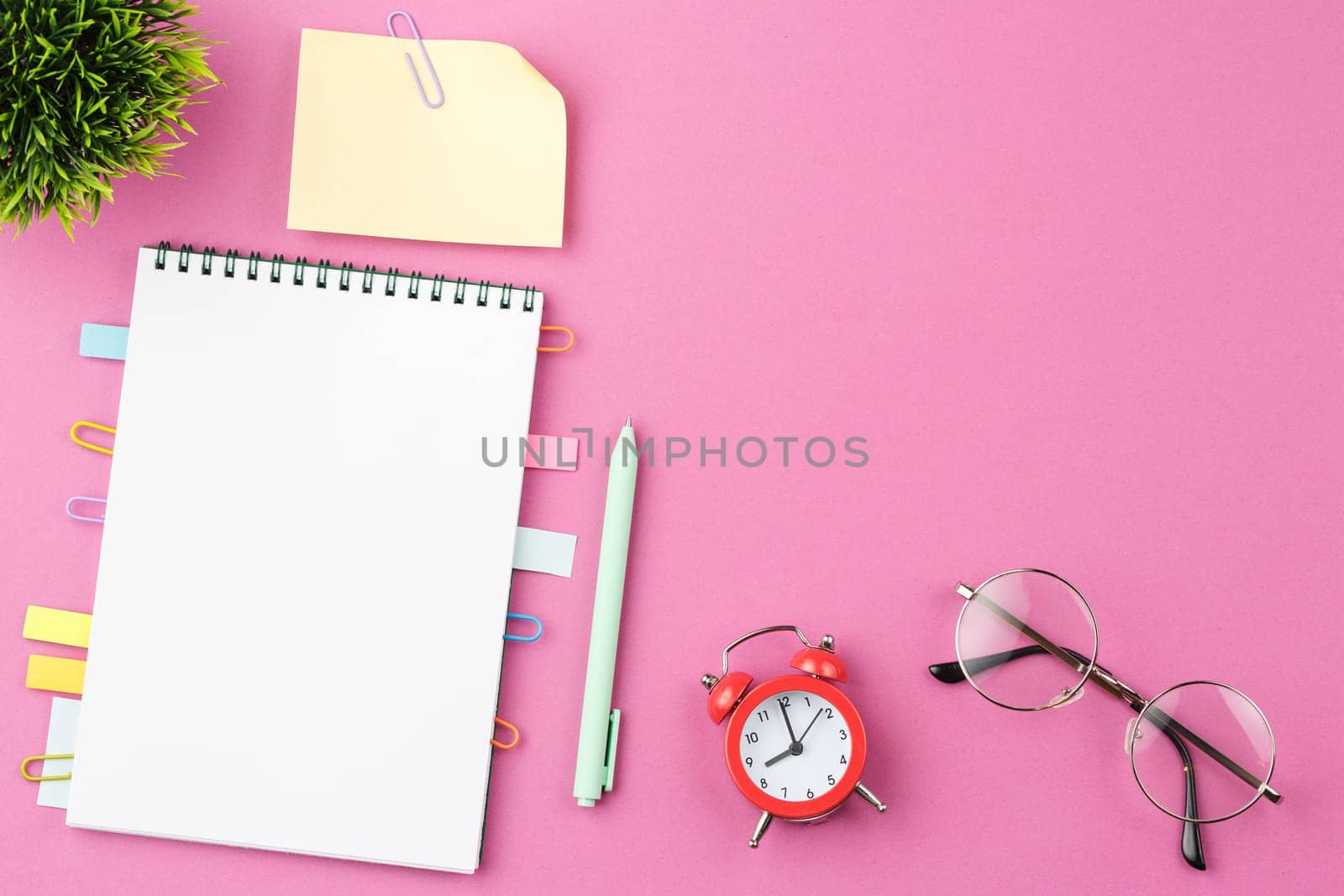 Open spiral notebook with bookmarks from paper clips and leaves for notes, pen, alarm clock, glasses and a flower in a pot on a pink background. Top view. Desktop office concept.