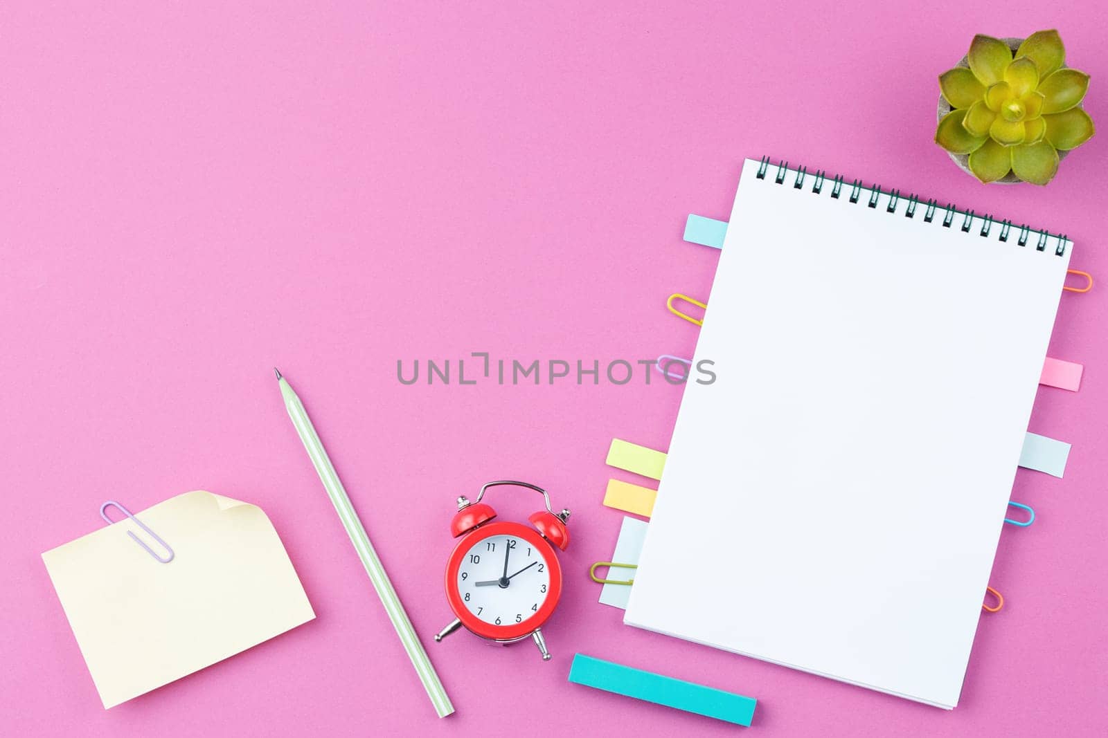 Open spiral notebook with bookmarks from paper clips and leaves for notes, pencile, rubber, alarm clock and a flower in a pot on pink background. Top view. Office desktop concept.