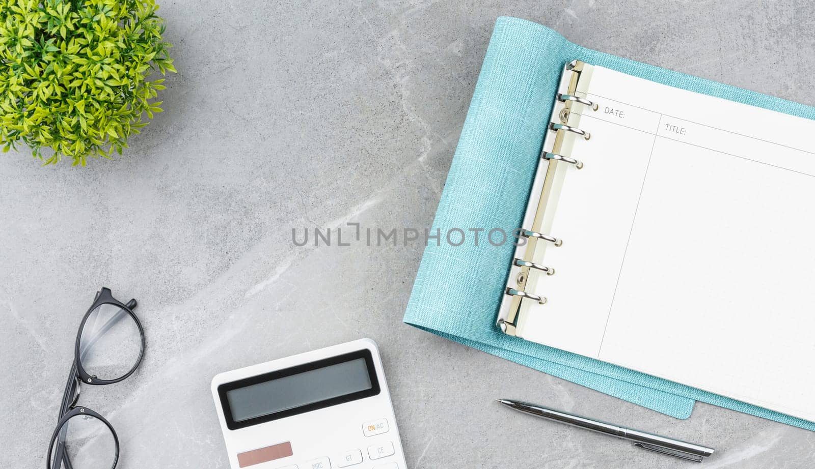 Spiral notebook in soft blue cover with metal pen, glasses, calculator and flower in a pot on gray marble background. Office desk concept. Top view.