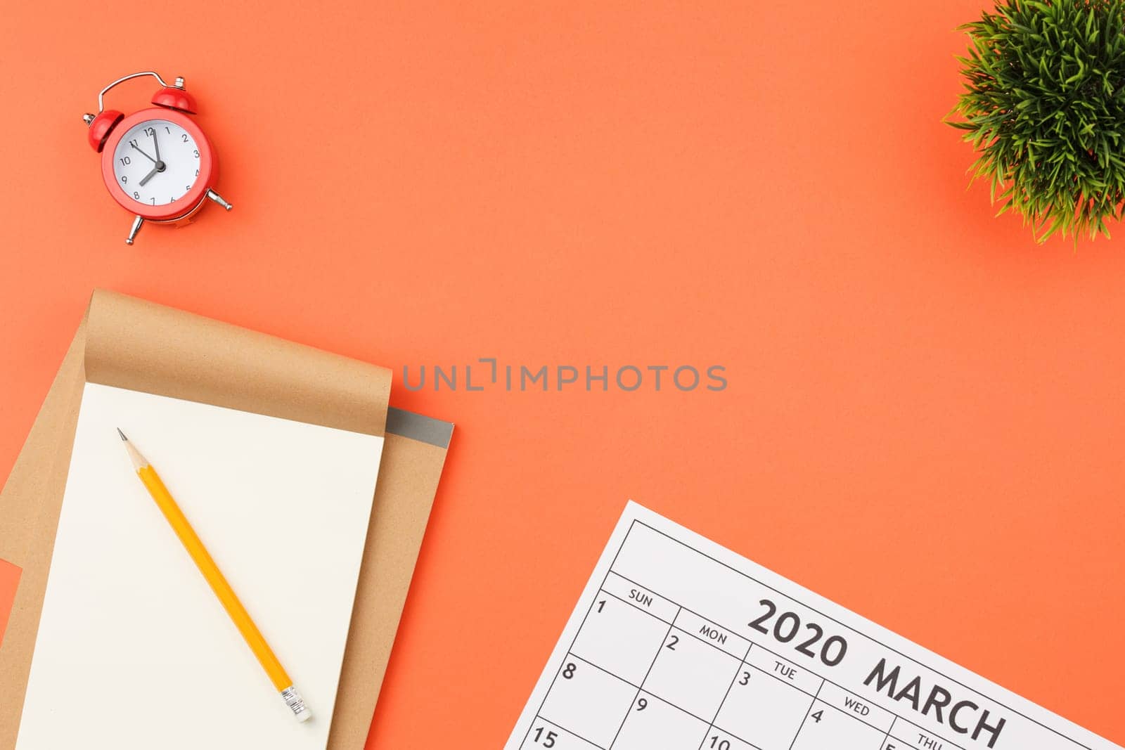 Craft notebook with white sheets, red alarm clock and pen, calendar sheet with dates and green leaves of a plant in a pot on an orange isolated background top view.