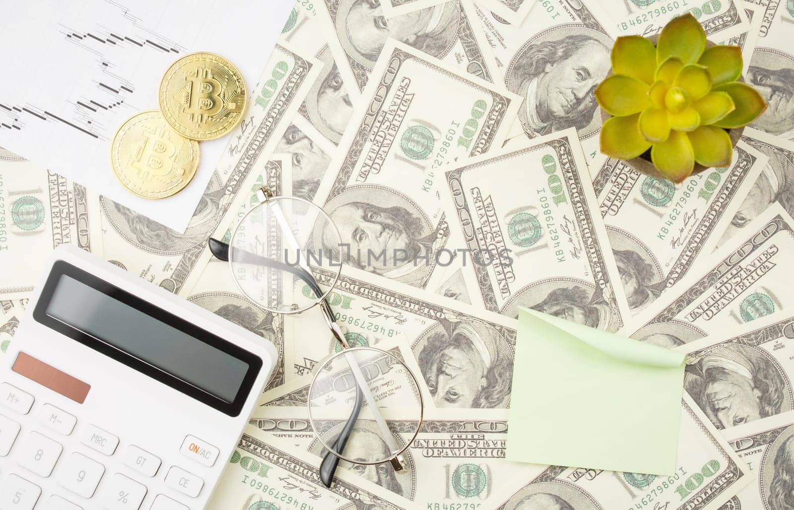 Bitcoin with lined hundred dollar bills background, stock chart, white calculator, glasses, note paper stick and spiral notebooks on brown background. Exchange trading and cryptocurrency concept.