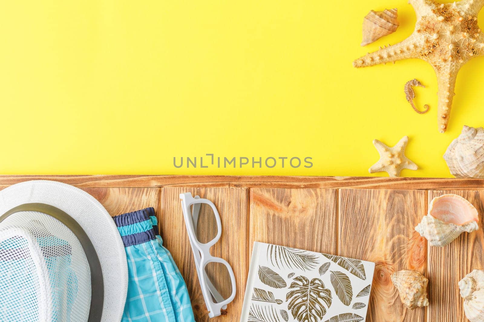 Beach hat with shorts and sunglasses on wooden background with yellow copy space. Flat lay. Summer vacation concept. Starfish and seashells. Top view.