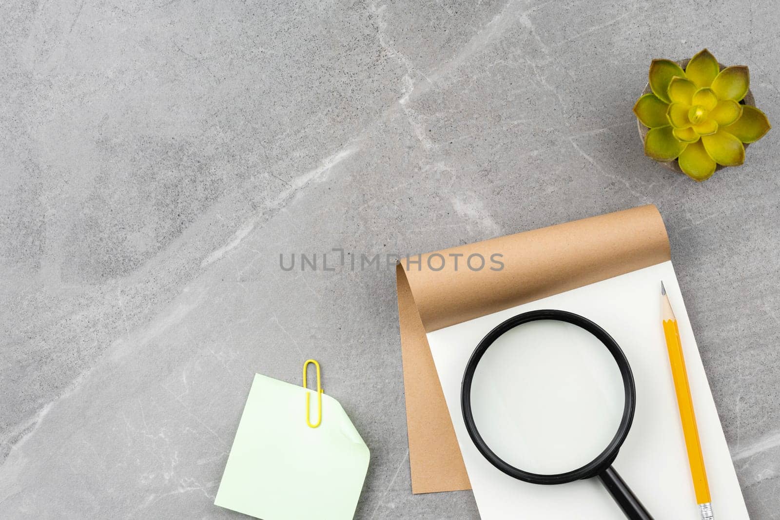 Open notebook, pencil, magnifying glass, notepaper and potted plant on gray marble background. Top view. Office desk concept.