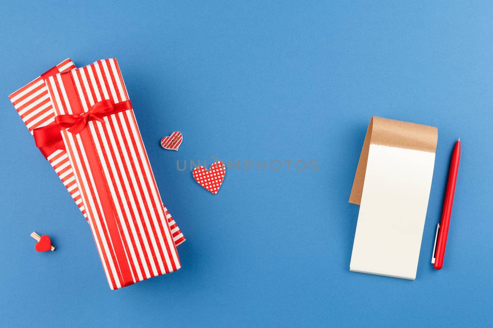 Present in striped paper with red satin bow on gray isolated background. Flat lay. Gift with hearts. Top view. Notepad with pen. Close up.